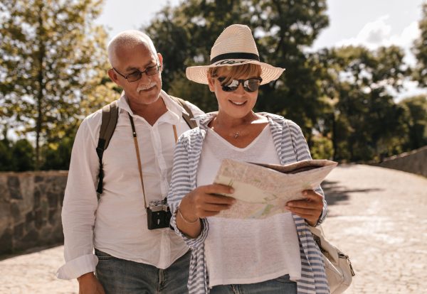 Blonde woman in white t-shirt, blue blouse, sunglasses and hat smiling and looking at map. Lady walks with mustachioed man in shirt with camera outdoor..